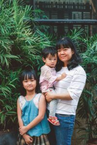 Woman holding a child, standing with another child in front of green foliage.
