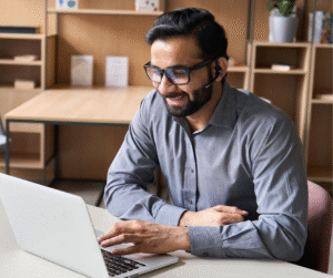 Man wearing glasses, working on a laptop at a desk, smiling.