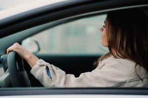 Woman driving a car, hands on steering wheel. She's wearing a light colored jacket. Side view.