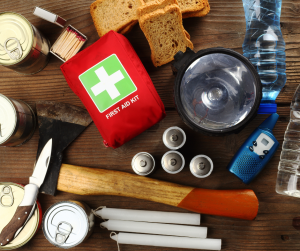 Emergency preparedness supplies on a wooden surface: first aid kit, canned food, flashlight, water, batteries, candles, ax, knife, matches, and bread.