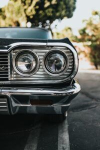 Classic car front end, chrome grill and headlights. Dark paint, outdoor setting.