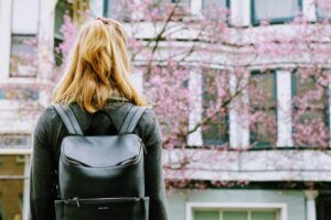 Woman with blonde hair wearing a black backpack, facing a building with pink blossoming trees.