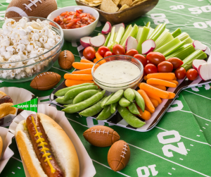 Football-themed snacks: hot dogs, popcorn, chips, vegetables with dip, salsa, and footballs on a green field.
