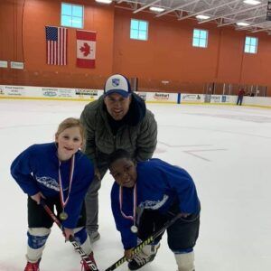 Three children and a coach pose on an ice rink, wearing blue hockey jerseys and medals.