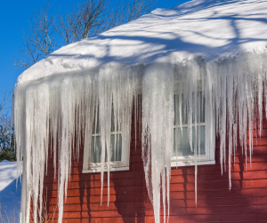 Red house with long icicles hanging from the snowy roof, two windows visible, bright blue sky.