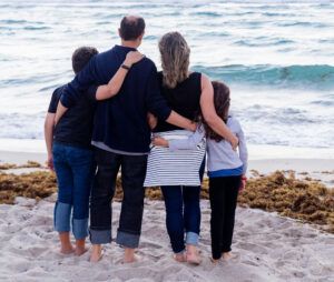 Family of four with arms around each other, facing the ocean. They stand on a beach with light brown sand.
