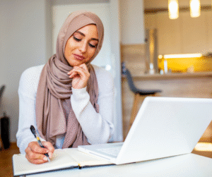 Woman in tan hijab, white top, writing in a notebook next to a laptop at a desk.