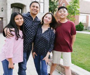 Family of four smiling, posing in front of a house. Adults in navy tops, children in pink and burgundy.