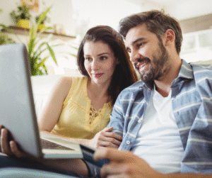 Couple shopping online, smiling at laptop screen, holding credit card on couch.