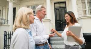 Real estate agent hands keys to a smiling couple in front of a white house with a black door.
