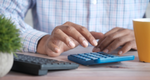 Hands using a blue calculator on a desk with a keyboard, plant, and mug.