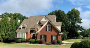 Brick house with brown roof, surrounded by green trees and bushes, against a blue sky.