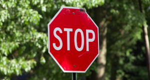 Red octagon stop sign with white text, in front of leafy green trees.
