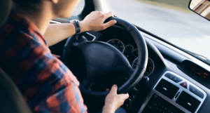 Person driving a car, hands on the steering wheel, wearing a plaid shirt.