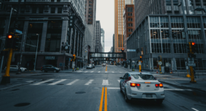 City street scene with car driving toward a pedestrian bridge, flanked by tall buildings, stoplights illuminated.
