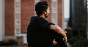 Man wearing glasses sits outdoors, looking toward buildings.