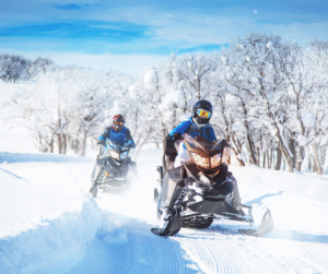 Two people riding snowmobiles on a snowy trail, under a bright blue sky.