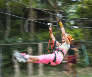Person zip-lining through a forest, arms raised, wearing helmet and safety gear.