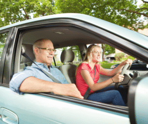Man in glasses sits next to a young person driving a car.