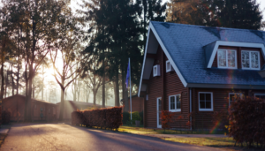 A wooden house with a blue roof sits next to a road, sunlight shines through trees.