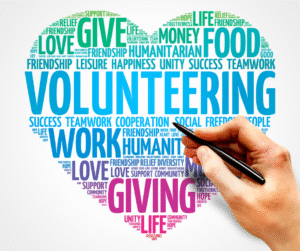 Hand holding a pen points to a heart-shaped word cloud with volunteering-related words.