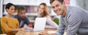 A smiling man leans toward the camera, near a group of blurred people and a laptop on a table.
