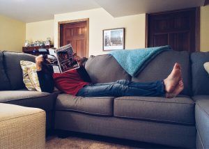 Person reading a magazine while lying on a gray couch in a room.