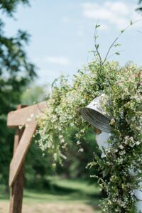 Wooden arch decorated with white flowers and a metal bucket, outdoors.