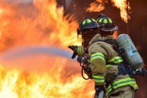 Two firefighters extinguishing a fire with a water hose; orange flames in the background.