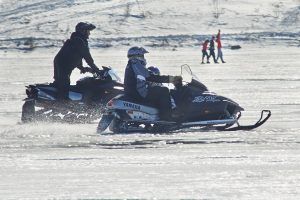 Two snowmobiles speeding across a frozen lake, with people in the background.