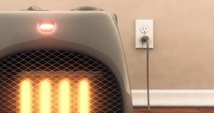 A gray space heater plugged into a wall outlet, glowing orange, indicating it is on.