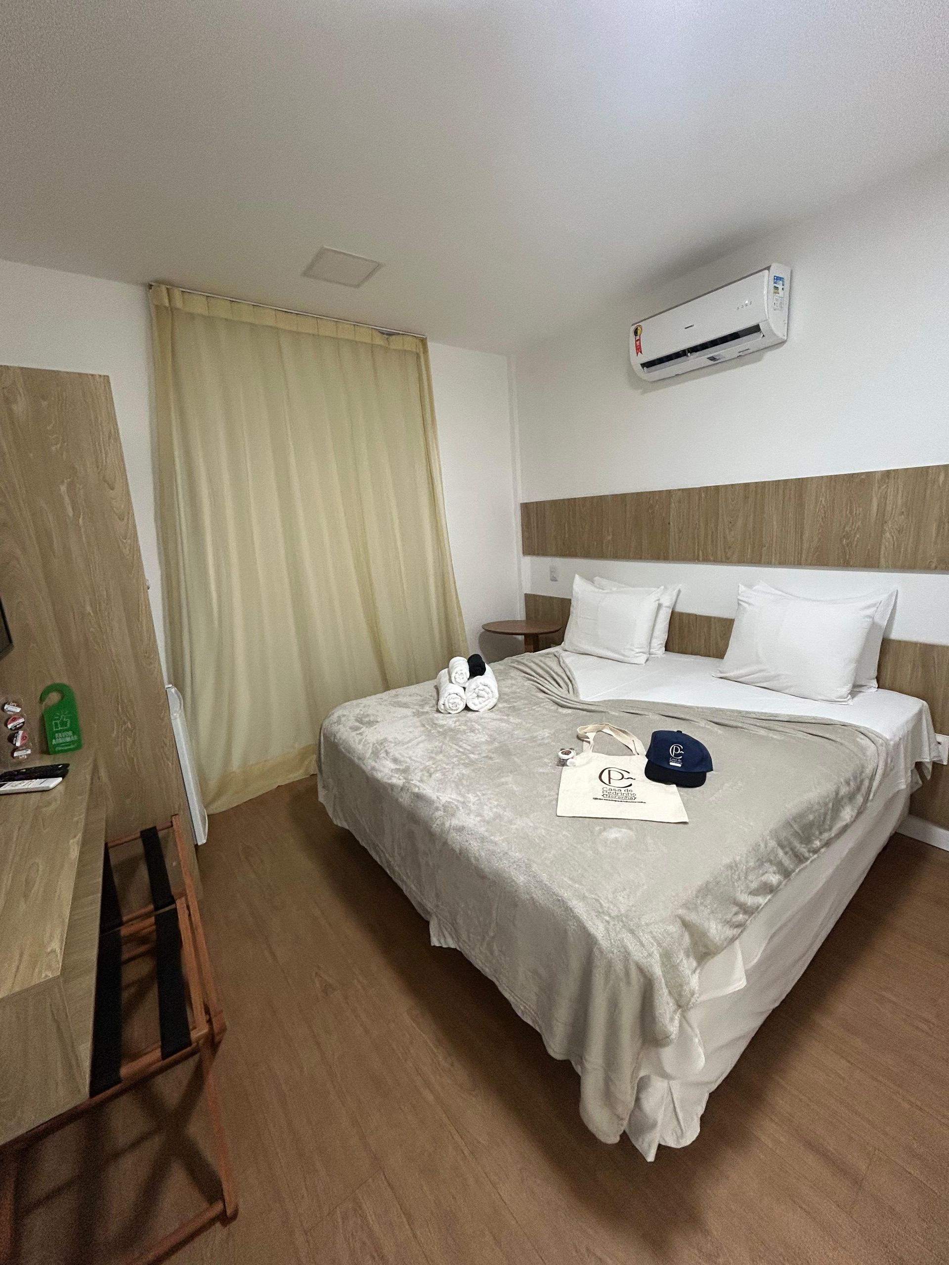 Quarto com cama coberta por um cobertor bege, travesseiros brancos, janela com cortinas translúcidas e uma escrivaninha.