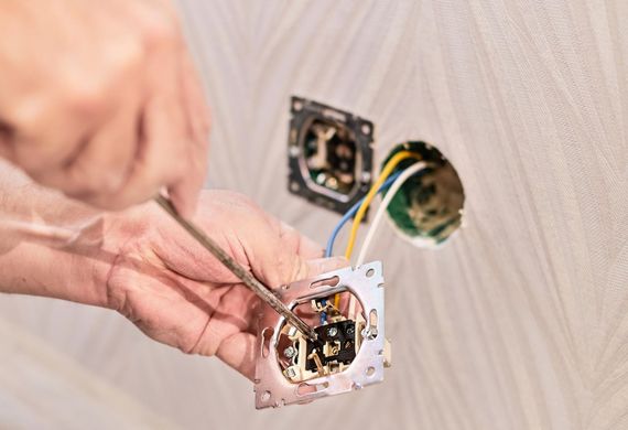 Hands wiring a wall outlet box with tools, exposed electrical wiring and a white textured wall