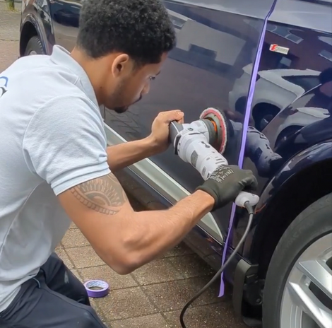 Iemand poetst een donkere autodeur met een polijstmachine; een paars snoer hangt er vlakbij.