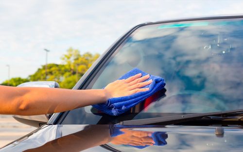 Handmatig een autoruit afvegen met een blauwe doek in fel daglicht.