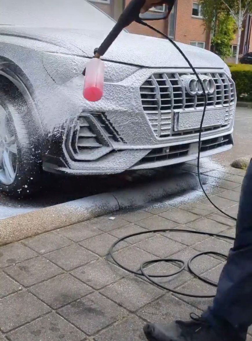 Een persoon reinigt een grijze Audi SUV op een oprit met een hogedrukreiniger en zeepschuim, terwijl er een rode spuitfles in de buurt staat.