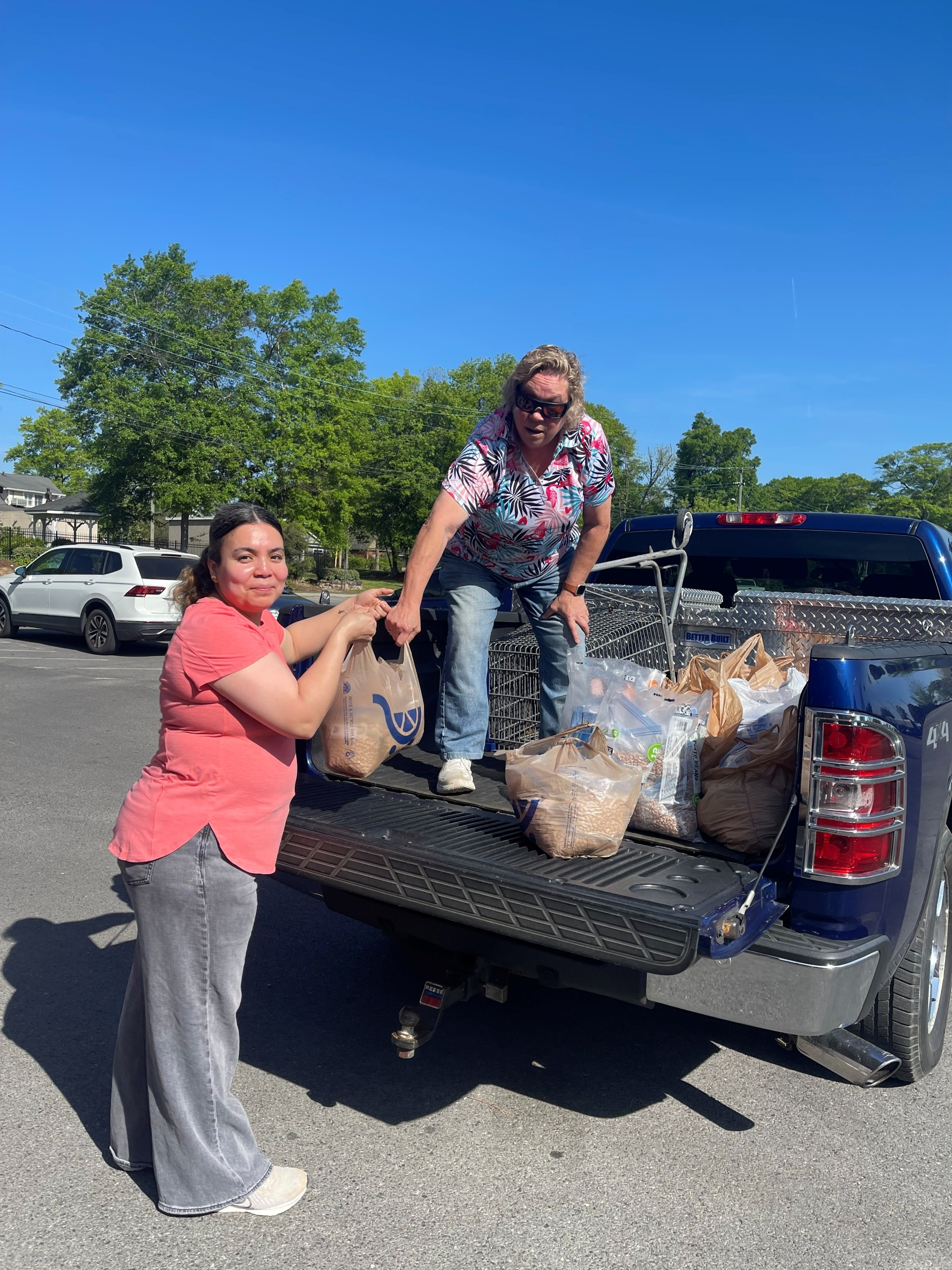 Two people loading bags into a black pickup truck in a parking lot on a sunny day.