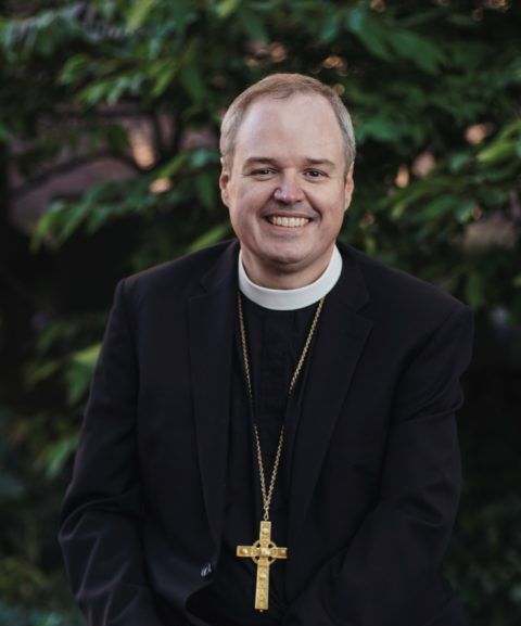 Smiling priest in a black suit with a gold cross pendant, seated outdoors against greenery