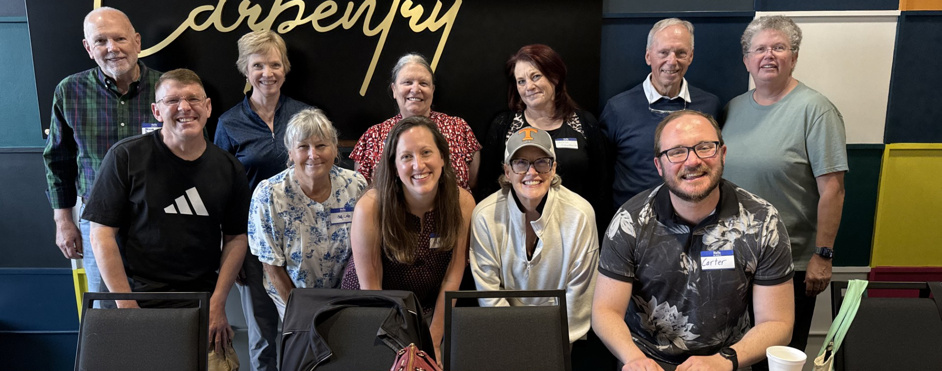 Group of people smiling in front of a decorative wall and black chairs in a classroom or meeting room.