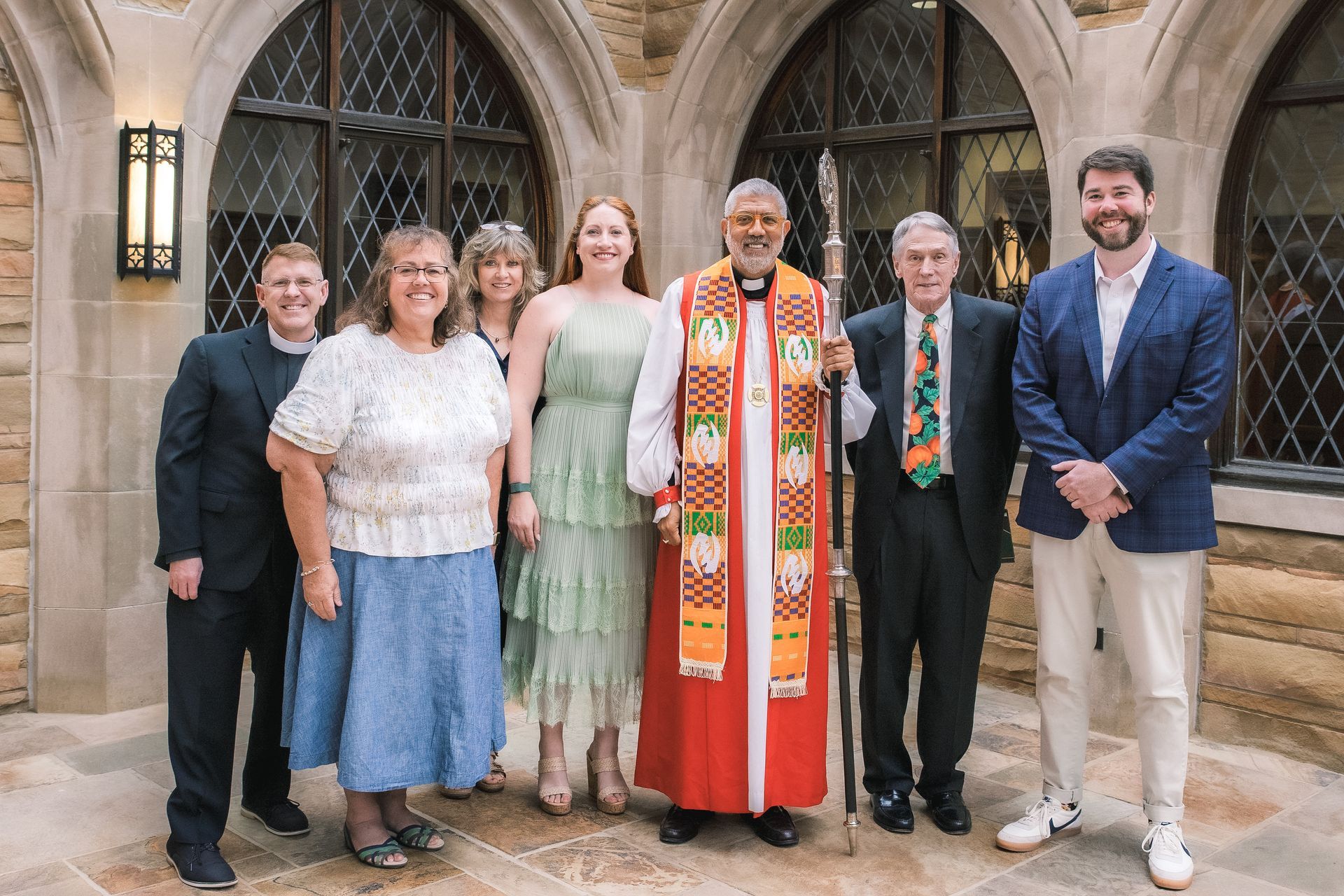 Six people posing on stone steps outside an ornate building, centered on two clergy in colorful robes.