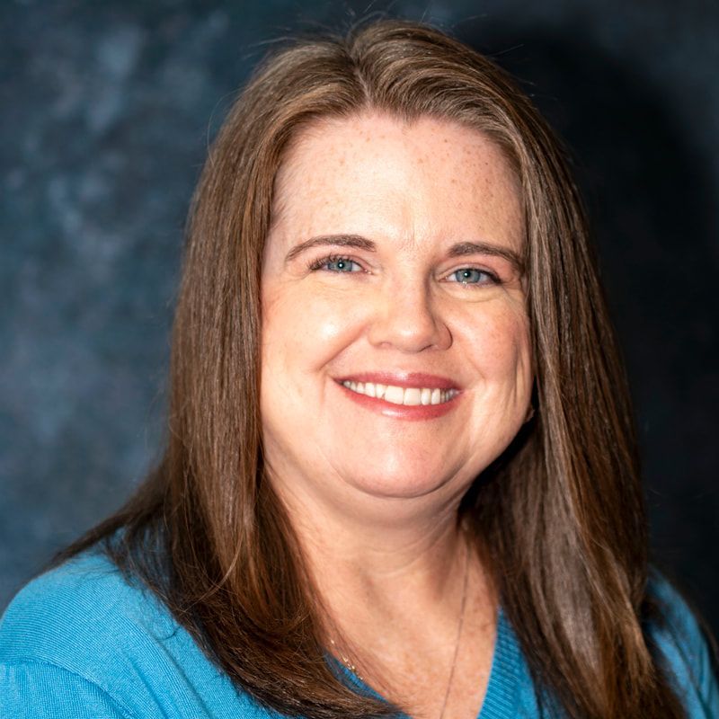 Smiling woman with straight brown hair wearing a blue top against a blue studio backdrop