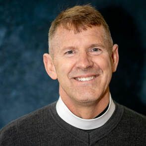 Smiling person in a dark clerical collar against a blue studio background
