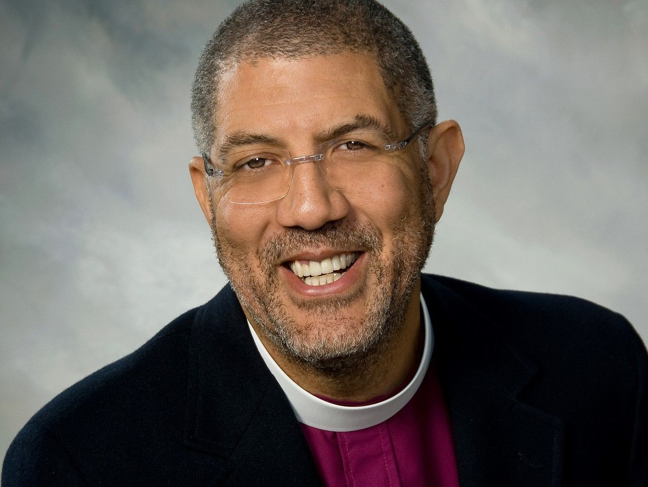 Smiling clergyman in black jacket and white collar against a gray background