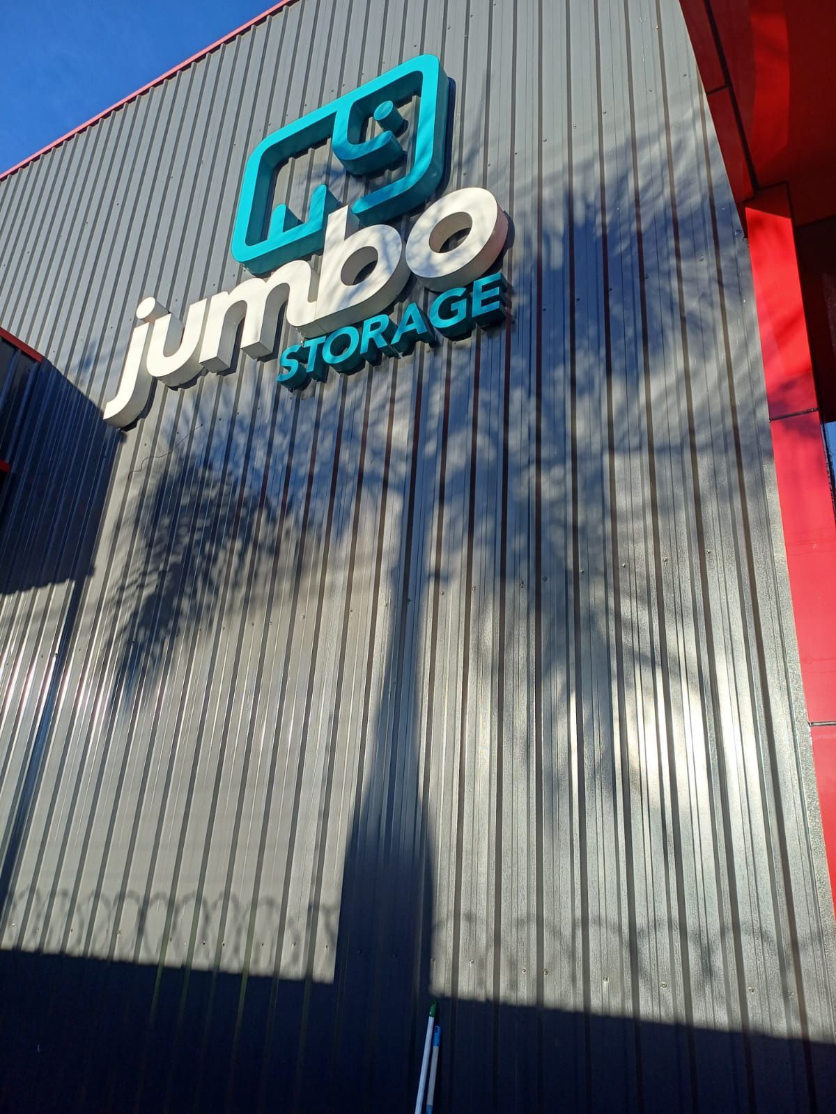 A placa da Jumbo Storage, com o logotipo de um elefante azul, está fixada em uma parede vertical de metal cinza, projetando uma longa sombra de palmeira.