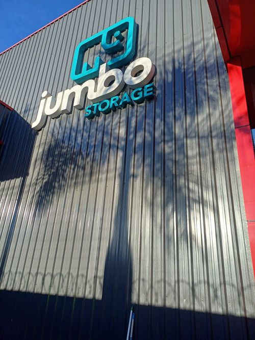 A placa da Jumbo Storage, com o logotipo de um elefante azul, está fixada em uma parede vertical de metal cinza, projetando uma longa sombra de palmeira.