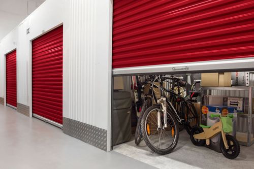 Um corredor com unidades de armazenamento com portas de enrolar vermelhas, uma delas parcialmente aberta revelando bicicletas e caixas embaladas no interior.