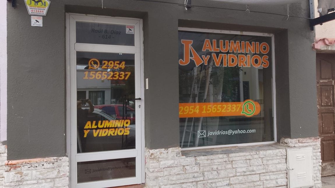 Fachada de “Aluminio y Vidrios” con fachada gris, puerta de cristal y letrero en la ventana.