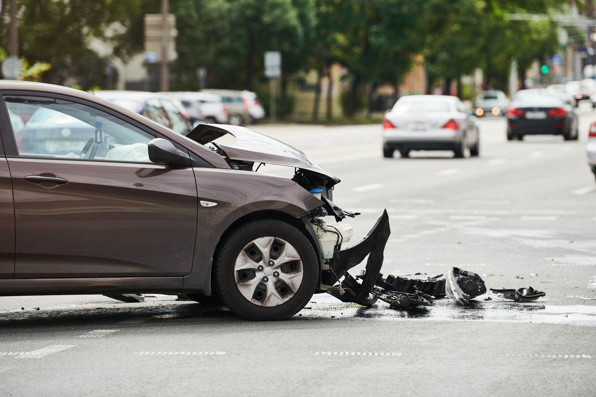 Damaged brown sedan after a front-end collision on a city street, with debris on the road.