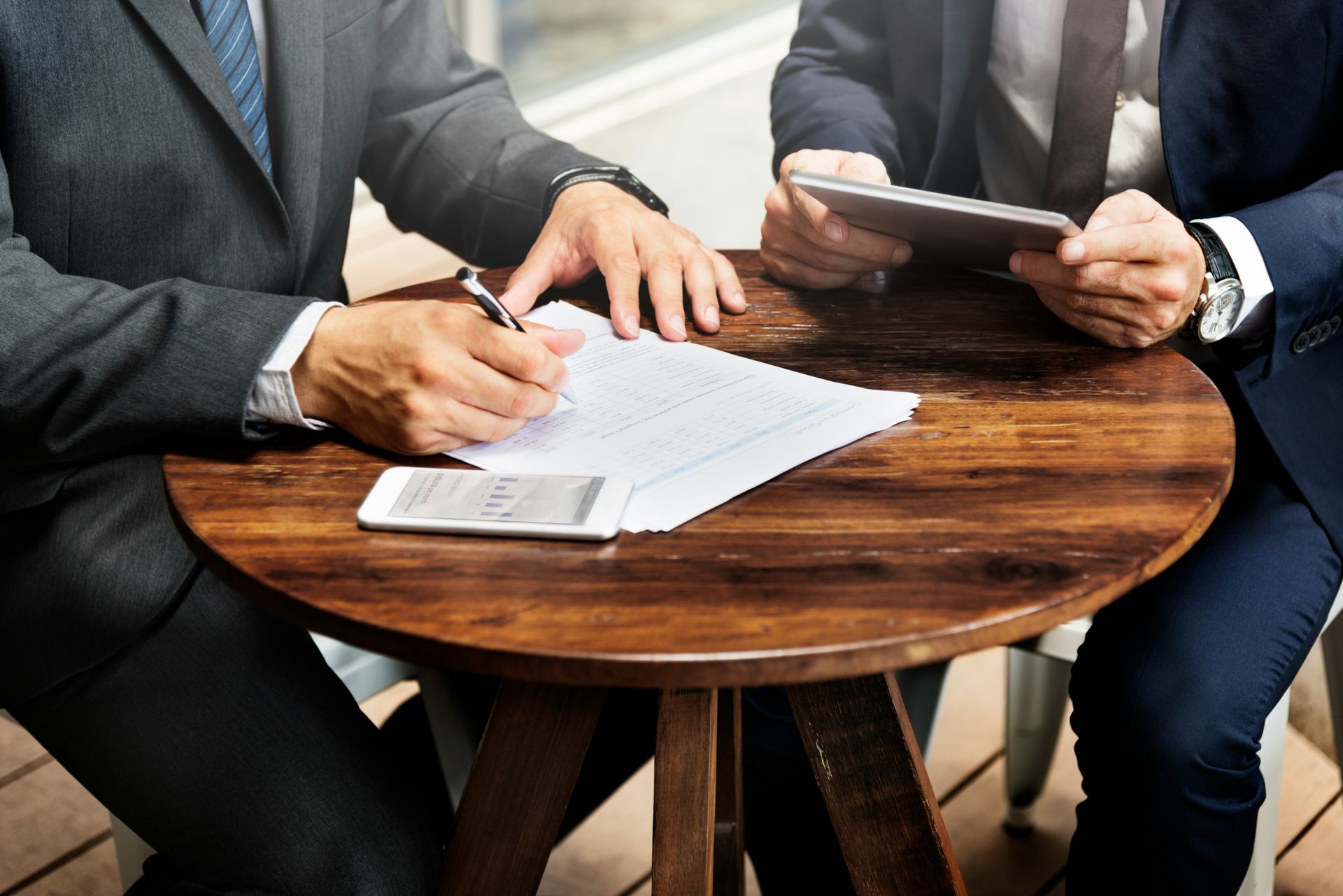 Two businesspeople in suits at a round table, one signing a document, the other holding a tablet.