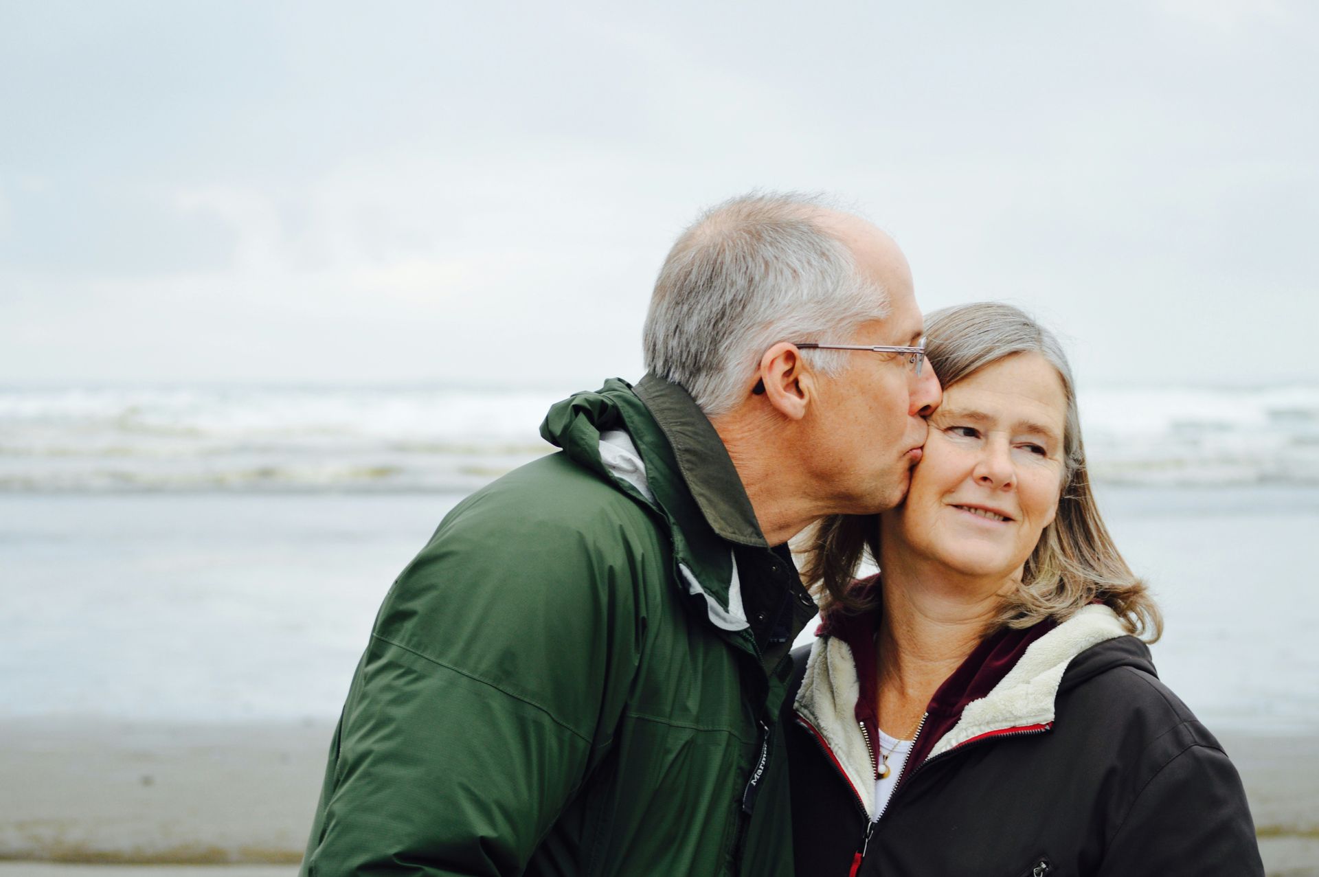 Man kissing woman's cheek on a beach; ocean in background. They wear jackets. Cloudy sky.